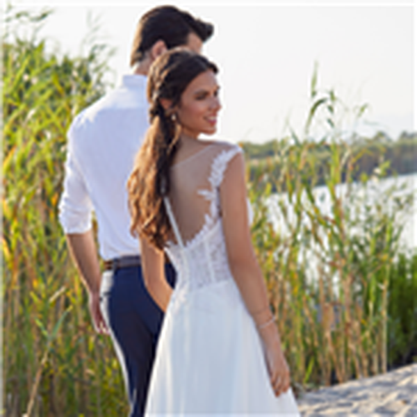 Thumbnail photo of the back of bride and groom models stood by tall reeds on a beach. Bride wears Ronald Joyce 18413, a white A-line wedding dress with an illusion necklace back